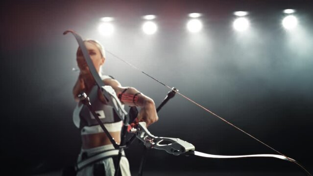 Cinematic Portrait of a Female Archer Holding a Modern Compound Bow, Aiming at a Target. Young Athlete Drawing and Releasing the Arrow in a Stadium Competition or in a Dark Studio for Advertising