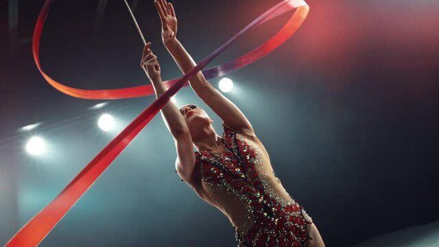 Portrait of a Beautiful Young Rhythmic Gymnast Performing a Dance with a Red Ribbon. Young Female Spinning the Ribbon in Striking Uniform on a Dark Stage. Cinematic Super Slow Motion Footage