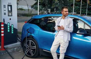 Standing with smartphone. Man with blue electric car on the charge station