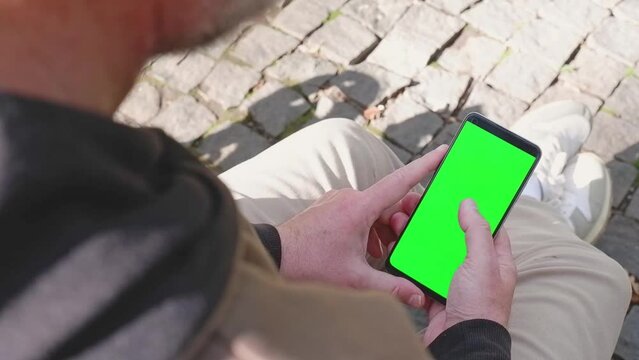 Close-up of a man's hand sitting in a park, holding his smartphone with a green screen and looking at it, scrolling, touching the screen. Back view. Layout.