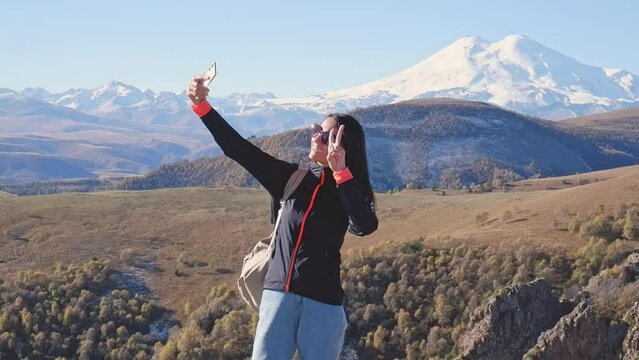 A female traveler with a backpack on her back on a hike in the Caucasus mountains. A happy young woman takes a photo of Mount Elbrus on her phone
