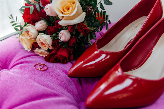 Red patent leather shoes of the bride near gold wedding rings and a bouquet of roses