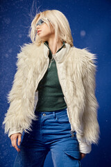 blonde woman in white faux fur jacket and sunglasses posing under falling snow on blue backdrop