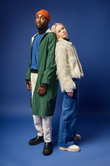 blonde woman leaning on african american man in winter attire, diverse young couple on blue backdrop