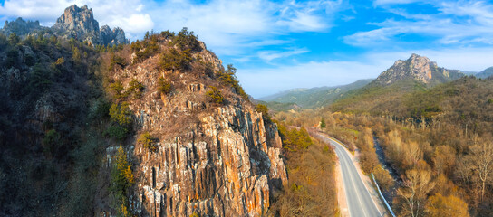 Mountain nature with highway. Rocky landscape view from quadcopter. Autumn nature. Mountains rise above road. Autumn rock with blue sky. High mountains with autumn trees. Picturesque landscape