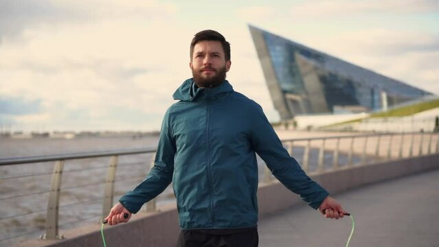 A hardy male athlete with a beard uses a jump rope intensive cardio training jumps on the spot and fitness equipment against the background of the city.