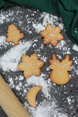 Black table with flour spread on it and freshly made homemade Christmas cookies. Tree, moon, reindeer, snow. A dark green kitchen towel and a dough rolling pin. Bakery cooking scene at home in winter.