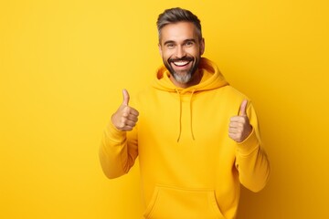 a studio portrait of a cheerful middle-aged man in a vivid bright yellow hoodie smiling and showing approval by holding a thumbs up on a seamless background