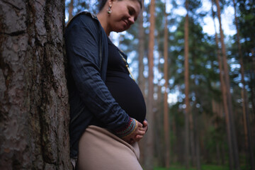 a pregnant woman stands leaning against a tree in the forest and looks at her tummy
