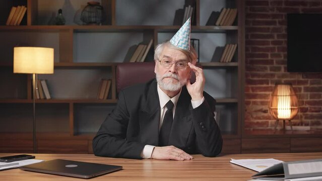 Portrait of serious older adult in formal clothes and party hat sitting at writing desk in modern office. Efficient business proprietor planning corporate anniversary with teammates in meeting room.
