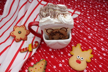 A Santa Claus-shaped mug with cream, stained with cocoa powder. A chocolate chip cookie peeks out of the cup. Homemade Christmas cookies. Red tablecloth with white stars and a striped kitchen towel. 
