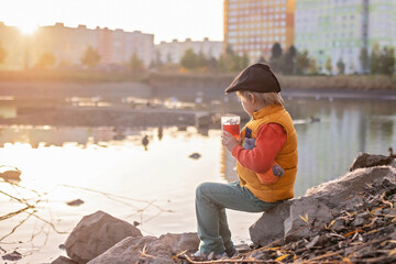 Beautiful blond preschool child, boy, sitting in the park in the morning sunrise, enjoying autumn weather, drinking tea and enjoying the sun