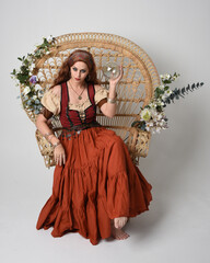 Full length portrait of beautiful red haired woman wearing a medieval maiden, fortune teller costume.  Sitting pose, holding a crystal orb. isolated on studio background.