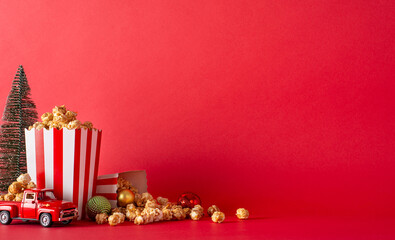 Create perfect Christmas movie night at home with delivered popcorn. Side-view image featuring popcorn boxes, balls, miniature vintage car, fir tree set against red wall, ideal for promotional content