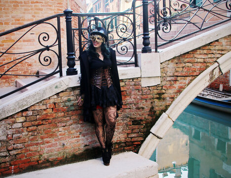 Steampunk girl in stockings, dress, corset, top hat, and retrofuturistic glasses poses in Venice canals. Her stylish attire and charming charisma captivate.