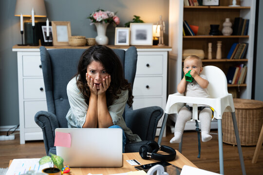 Young stressed single mother freelance worker business woman small company owner stressed and frustrated working from home office maternity leave while baby sitting next to her playing in baby chair