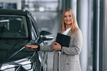 Black colored document in hands. Woman in formal clothes is in the car dealership