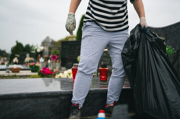 Professional gravesite care. Big autumn cemetery cleaning before the All Saints Day. Unrecognizable worker with big plastic bag and protective gloves collecting garbage.