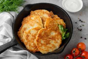 Potato pancakes, latkes or draniki with fresh herbs and sour cream in a cast iron pan on a gray concrete background. Top view, copy space.