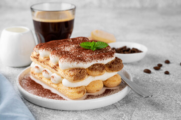 Tradition italian layered dessert tiramisu with mascarpone cream and biscuits on a white plate with cup of coffee on a gray concrete background.