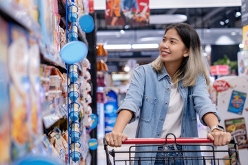 Happy smiling Asian woman wearing jeans with shopping cart in supermarket department. She is...
