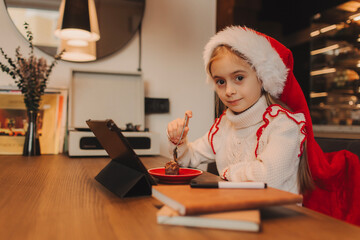 Happy girl enjoys the Christmas holidays, eating a chocolate dessert while looking at a tablet iPad...