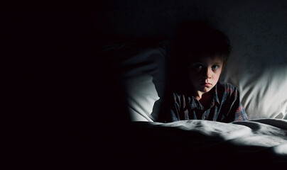 Small child boy lies in bed in dark night and covers his face with his hands in fear, afraid of nightmares and terrible dreams in children. Concept of horror and domestic violence - obrazy, fototapety, plakaty