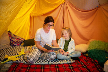 A son and his beautiful mother wearing glasses are reading a book together and having the best time bonding in an indoor comfy tent
