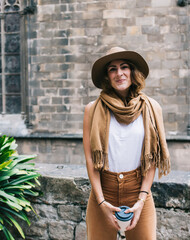 Stylish woman with disposable cup of coffee standing next to old stone building