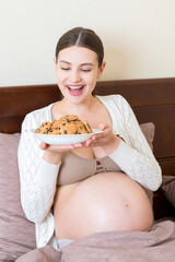 Pregnant woman enjoys eating cookies resting in bed at home. Unhealthy sweet pastry during pregnancy concept