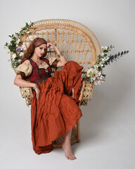 Full length portrait of beautiful red haired woman wearing a medieval maiden, fortune teller costume.  Sitting pose, with gestural hands reaching out. isolated on studio background.