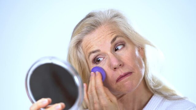 Closeup of mature middle aged woman 50s, 60s, applying natural makeup powder foundation with cosmetic sponge on clean face skin smoothing wrinkles, looking in mirror beauty routine white background.