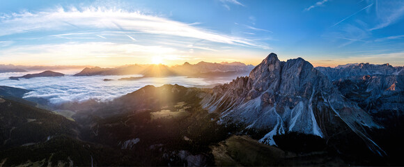 Sunrise over the Sass de Puta mountain peak at Passo delle Erbe pass against the Dolomite peaks in the background, inverse cloud cover in the valley, sun rays. Aerial drone mountainscape panorama.