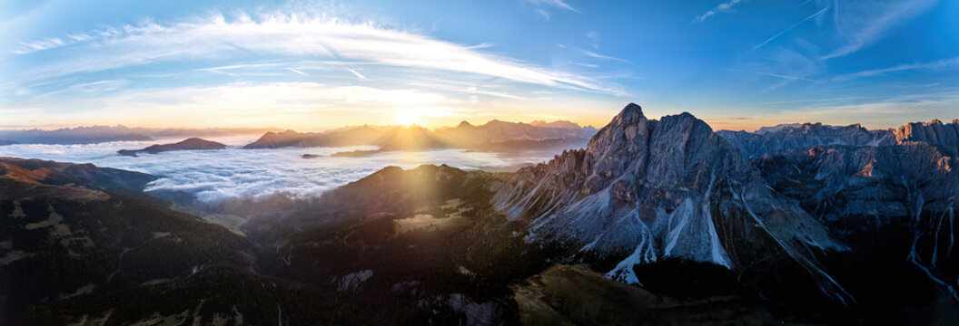 Sunrise over the Sass de Puta mountain peak at Passo delle Erbe pass against the Dolomite peaks in the background, inverse cloud cover in the valley, sun rays. Aerial drone mountainscape panorama.