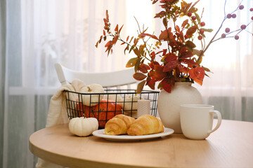 Autumn home interior. Two croissans on the plate, pumpkins in basket, vase with autumn leaves on...