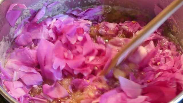 Cooking in water of peony flowers petals in metal pot - homemade peony jam, adding pink petals and mixing with a wooden spoon. Topics: natural vitamins, natural remedy for allergies, health
