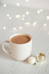 A white cup with hot coffee or tea on a white wooden table with Christmas lights.