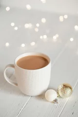  A white cup with hot coffee or tea on a white wooden table with Christmas lights. © Anna