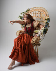 Full length portrait of beautiful red haired woman wearing a medieval maiden, fortune teller costume.  Sitting pose, with gestural hands reaching out. isolated on studio background.