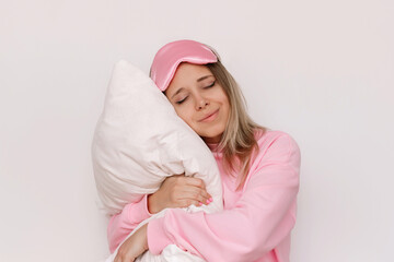 A young sleepy blonde woman in a pink pajamas and sleep mask with her eyes closed hugs a pillow...