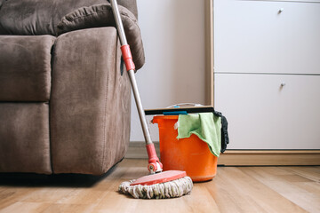 Plastic bucket with cleaning supplies and mopping stick near couch in home