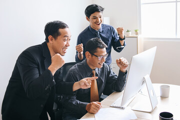 Group of Asian businessman looking at computer screen and screaming with joy, clenching fists celebrating successful business project.