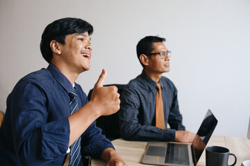 Young Asian smiling businessman showing thumbs up while sitting by senior male colleague in...