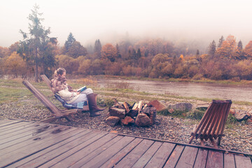Young woman wrapped in a cozy scarf. Warm colors of autumn. Vacation outside the city. Away from civilization. Reading a book wrapped in a plaid. Vacation in nature. Autumn foliage. 