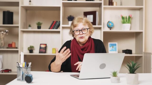 older woman in glasses sits at the table with a laptop, scolding while waving her hand and talking, looking at the camera