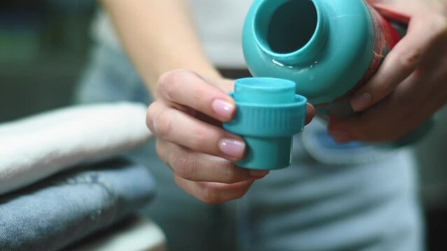 Close up of female hands pouring liquid laundry detergent into cap on table with stack clothes, blur background