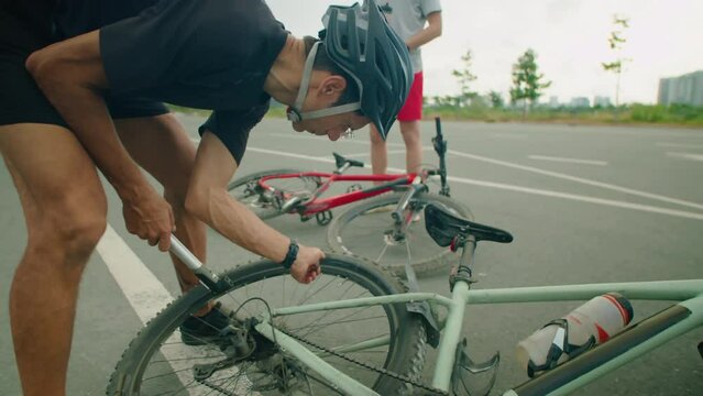 Man in cycling helmet inflating bike tire with pump as his son drinking water in background while having stop on road