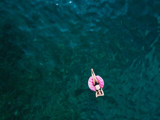 Aerial drone view of young woman with open arms in swimsuit lying and relaxing on big pink donut float swims on water surface with transparent bottom. Summer holiday vacation at exotic resort