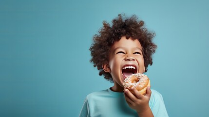 A little boy with a smile is eating a donut on a blue background wall. The child is having a good time with the donut. It's a fun time to have sweet food at home. - obrazy, fototapety, plakaty