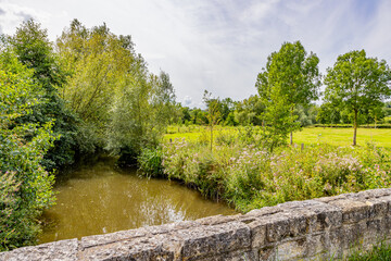 River Geul among wild plants and leafy trees seen from stone bridge, plain with green grass against gray cloud-covered sky in background, cloudy day at Terpoorterweg, Epen, South Limburg, Netherlands - Powered by Adobe